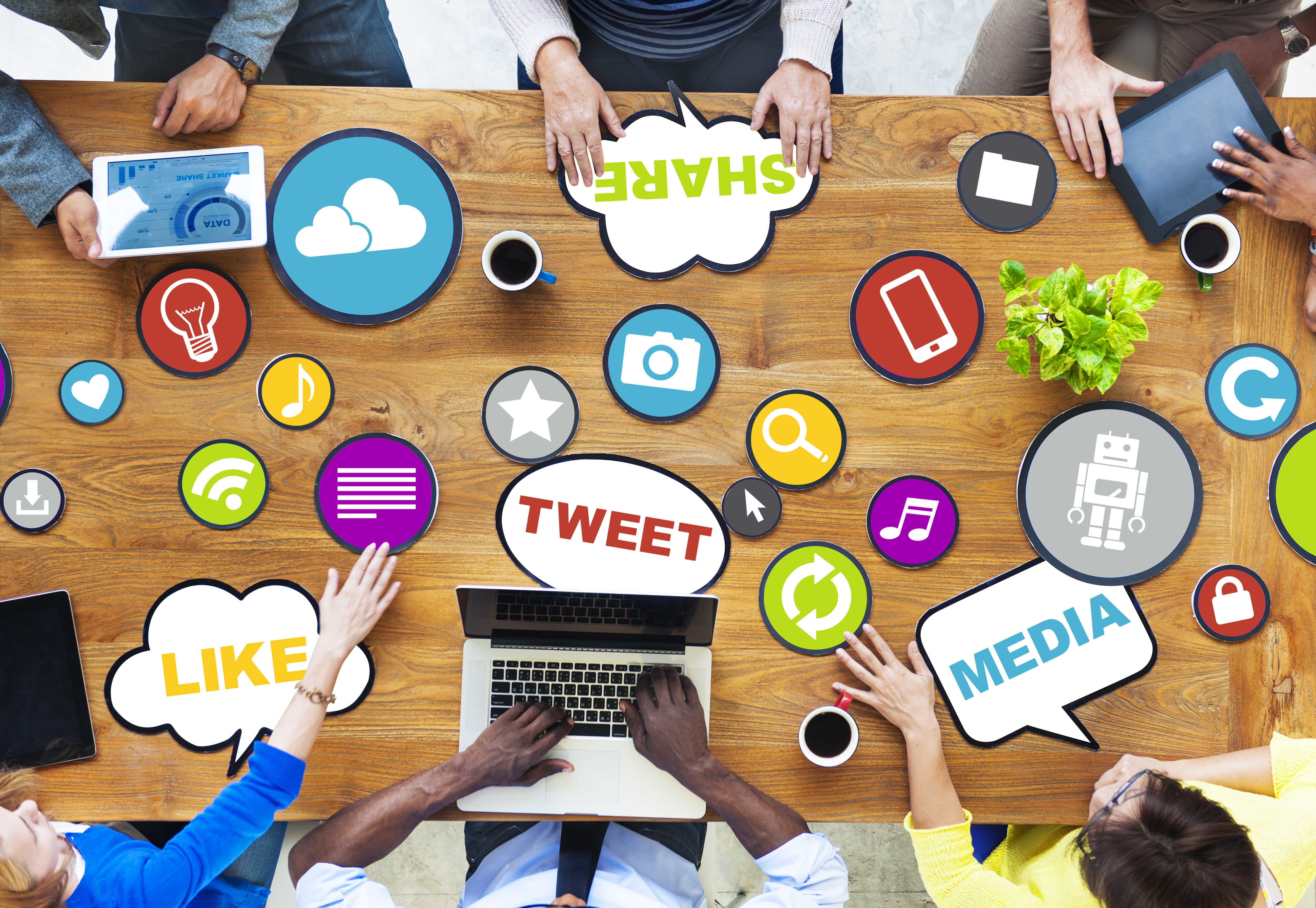 Large wooden table with 6 people holding different devices and icons scattered all around the table.